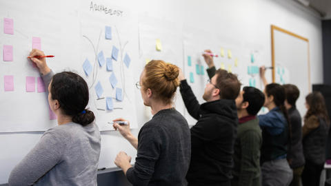 Pedagogy fellows write on sticky notes on chart paper stuck to the wall during a meeting.