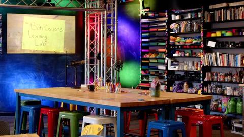 The Bok Center Learning Lab - a space with a large butcher block table, colorful metal stools around it, and bookshelves full of colorful paper and craft supplies in the background. A projector screen shows a piece of yellow paper reading "Bok Center Learning Lab."