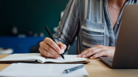 person writing on paper next to computer