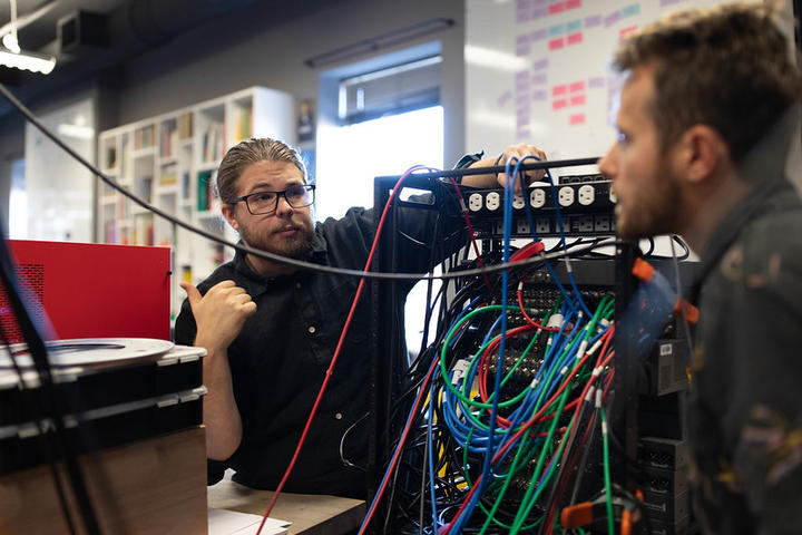 Casey talks with a Media Production Assistant over a piece of equipment with a tangle of colorful wires in the Learning Lab Studio.