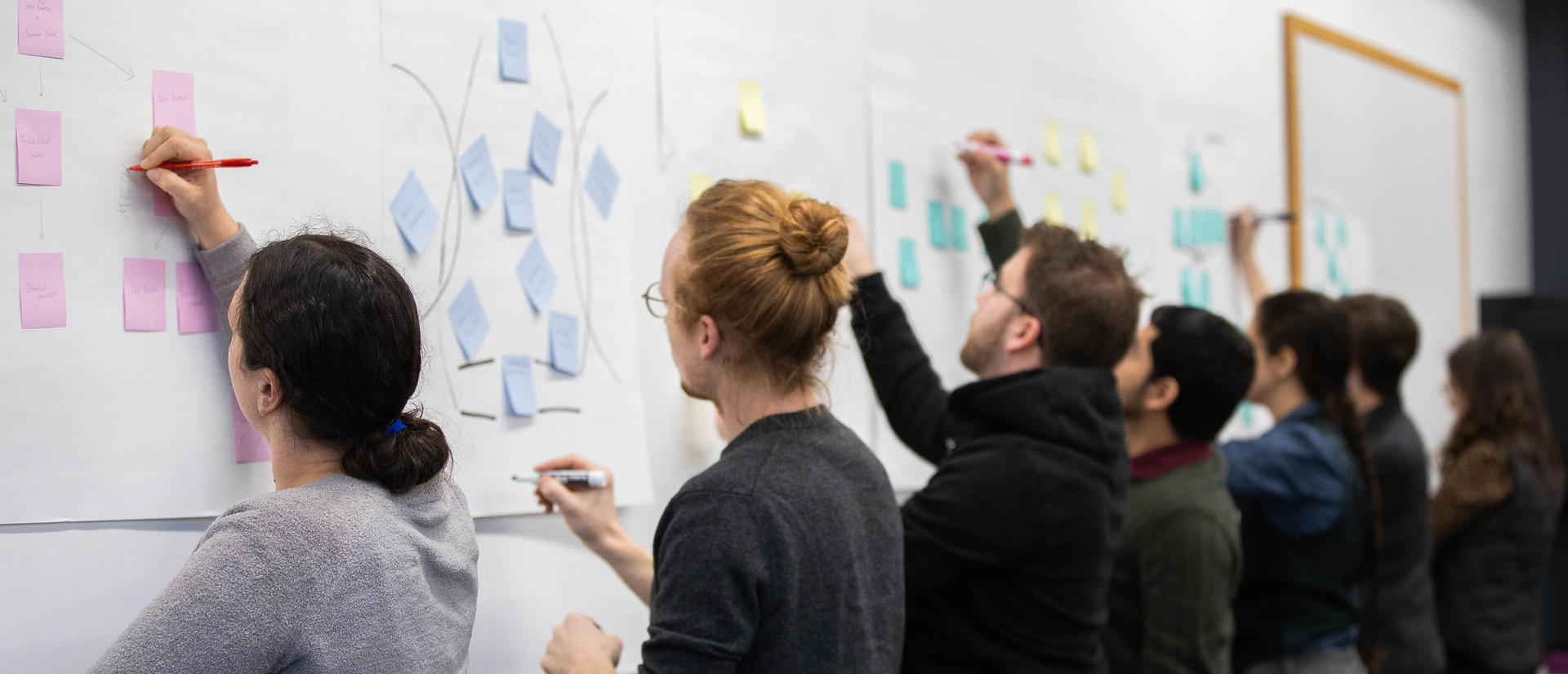 Pedagogy fellows write on sticky notes on chart paper stuck to the wall during a meeting.